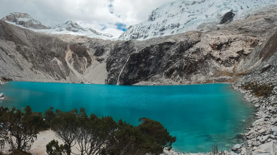 Turquoise lake below snow-capped mountains with waterfall.
